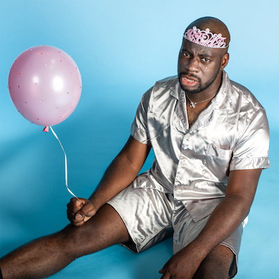 Album art for BRAT: NNAMDÏ, dressed in a shiny silver shirt and shorts, wears a pink tiara and holds a pink balloon while sitting in front of a blue backdrop.