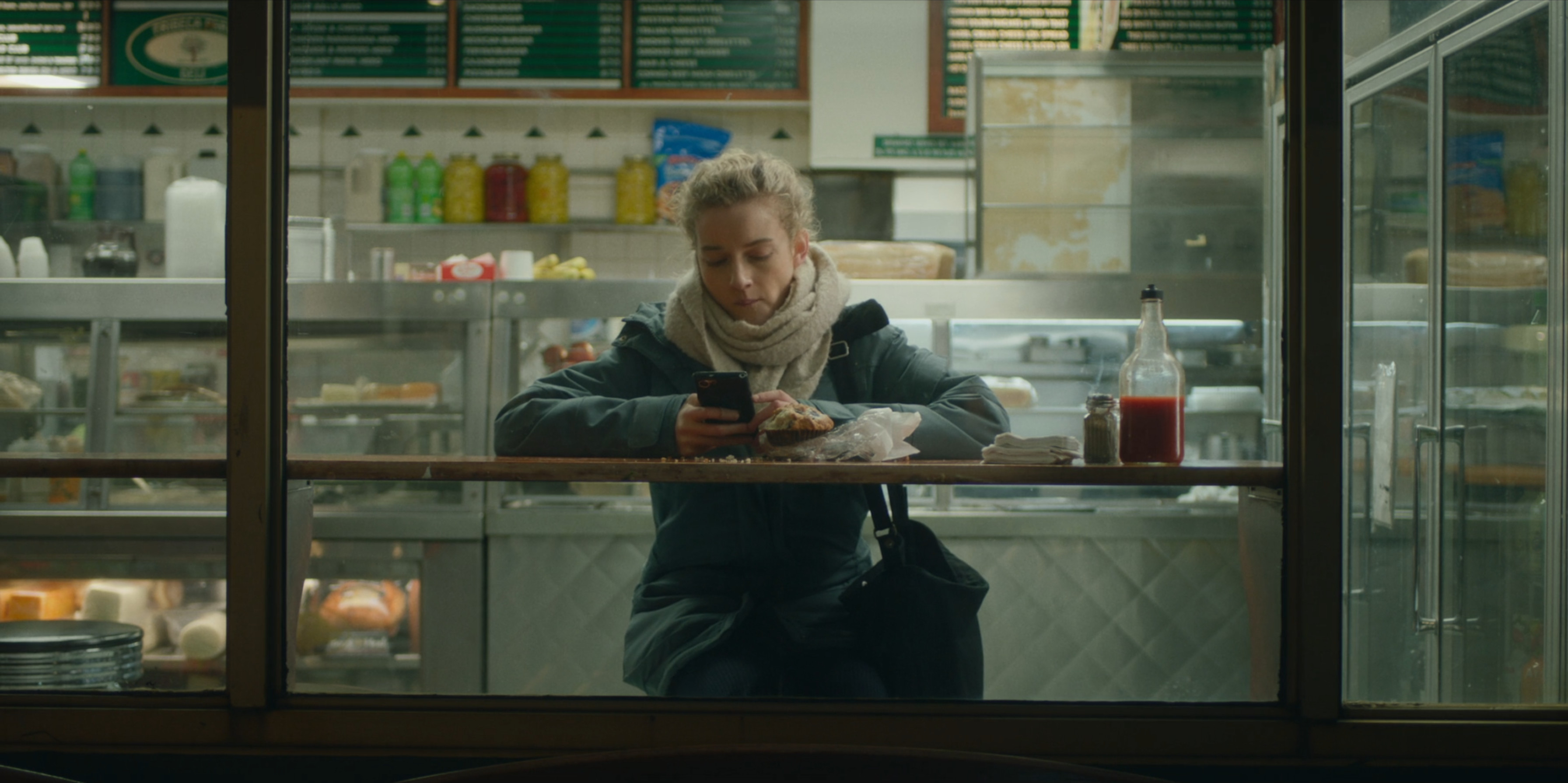 Through a window, we see a woman sitting in a cafe, facing towards us but looking at her phone.